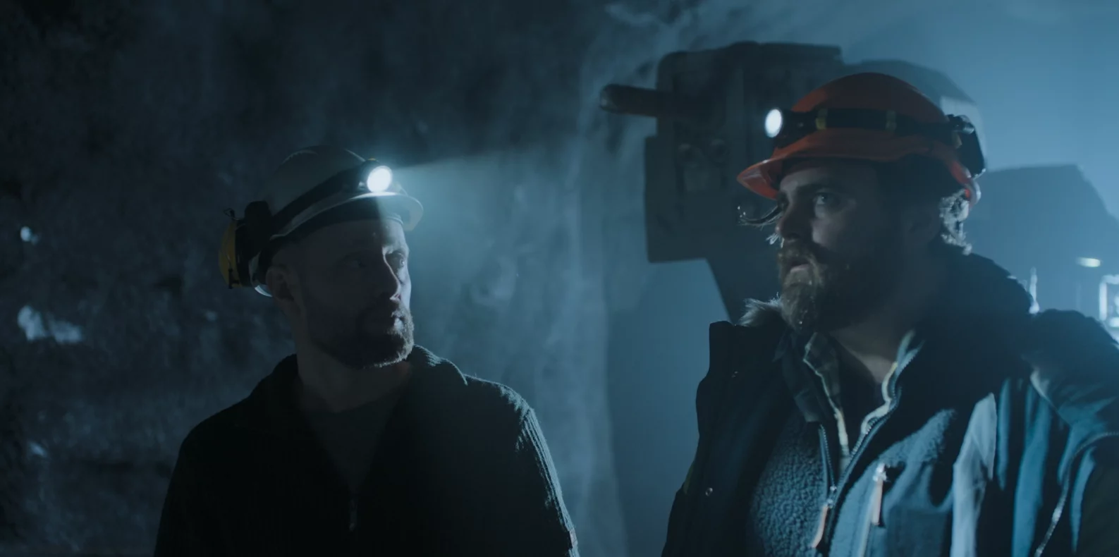 Zwei Männer mit Schutzhelmen und Stirnlampen stehen in einem Bergwerk. Die Szenerie ist düster. Der Linke blickt den rechten an, welcher an der Kamera vorbeiblickt.